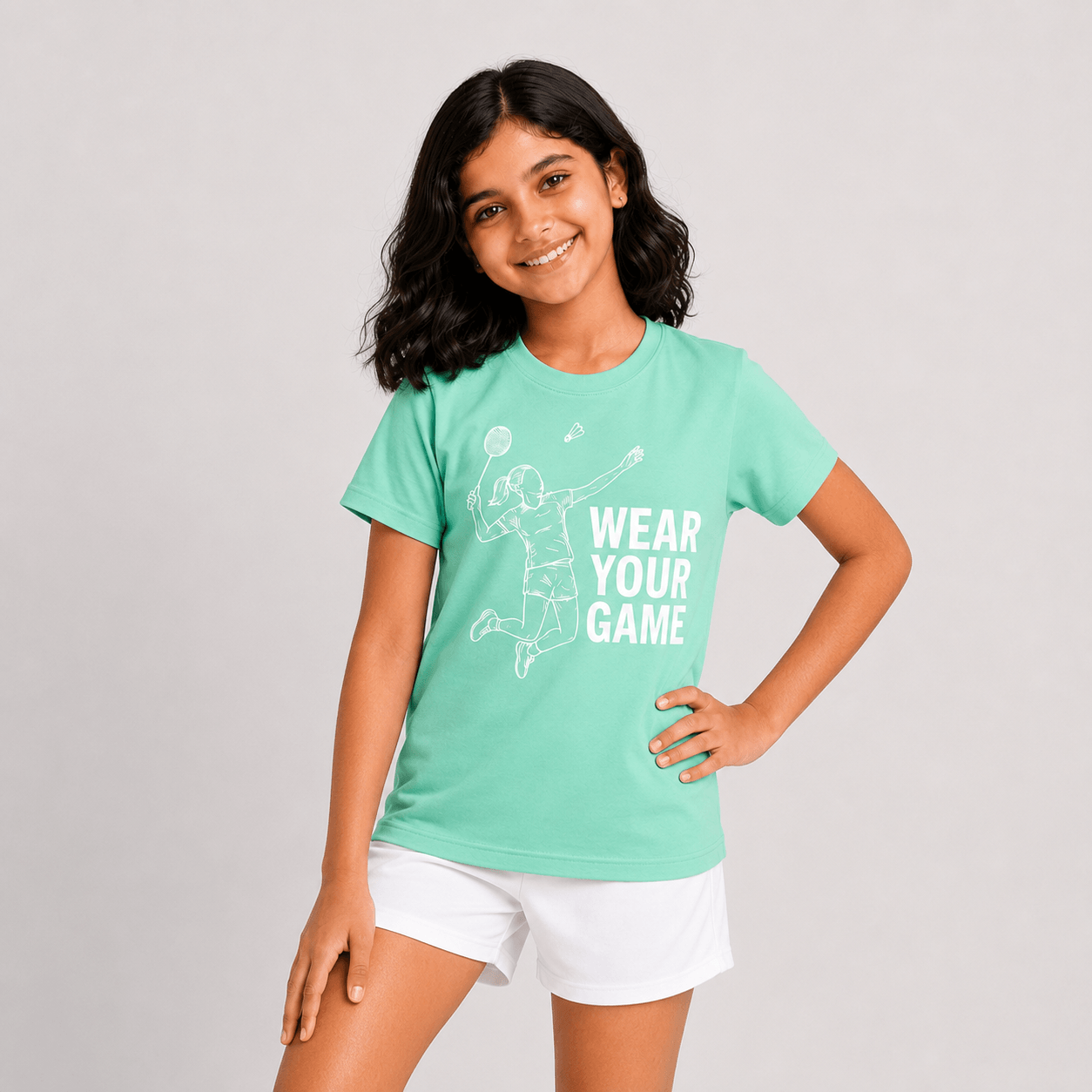 A young girl with dark hair is smiling and posing in a mint green t-shirt with a white graphic that reads 'WEAR YOUR GAME' and white athletic shorts, set against a plain light gray studio background.