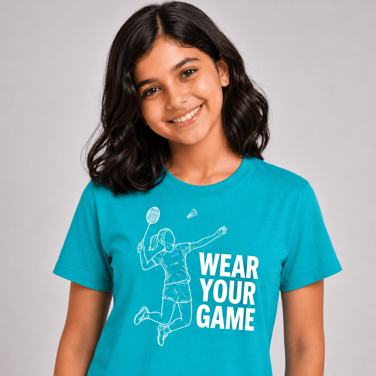 A smiling young woman with dark hair wearing a bright turquoise blue t-shirt with a white graphic of a badminton player and the text 'WEAR YOUR GAME'.