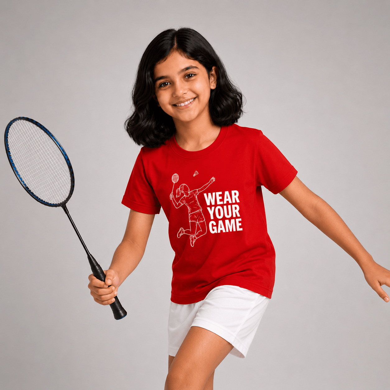 A young girl smiling and holding a badminton racket, wearing a red graphic t-shirt that says 'WEAR YOUR GAME' in white text with a white line drawing of a player, paired with white shorts, against a solid gray studio background.