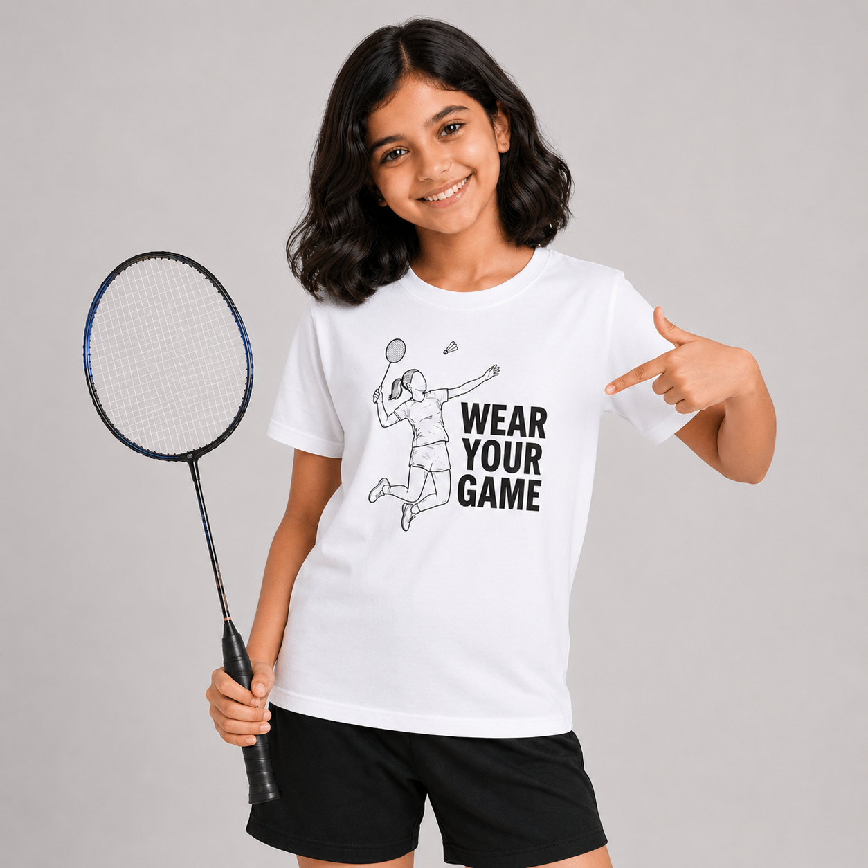 A young girl with dark hair smiling and pointing to a white graphic t-shirt. The t-shirt features a black line art illustration of a badminton player next to the text 'WEAR YOUR GAME'.