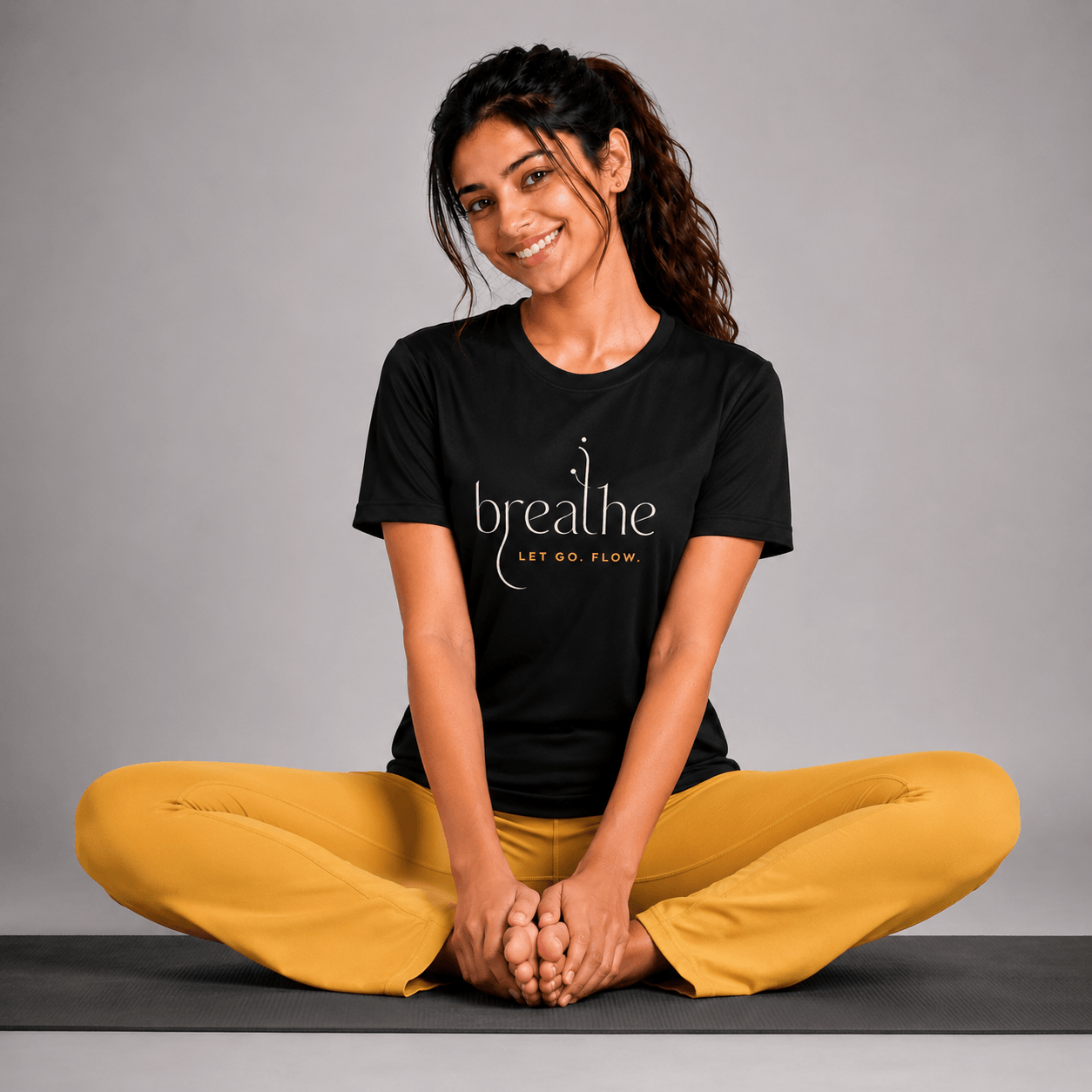 Woman sitting on a yoga mat wearing a black t-shirt with 'breathe' text and yellow pants against a gray background.