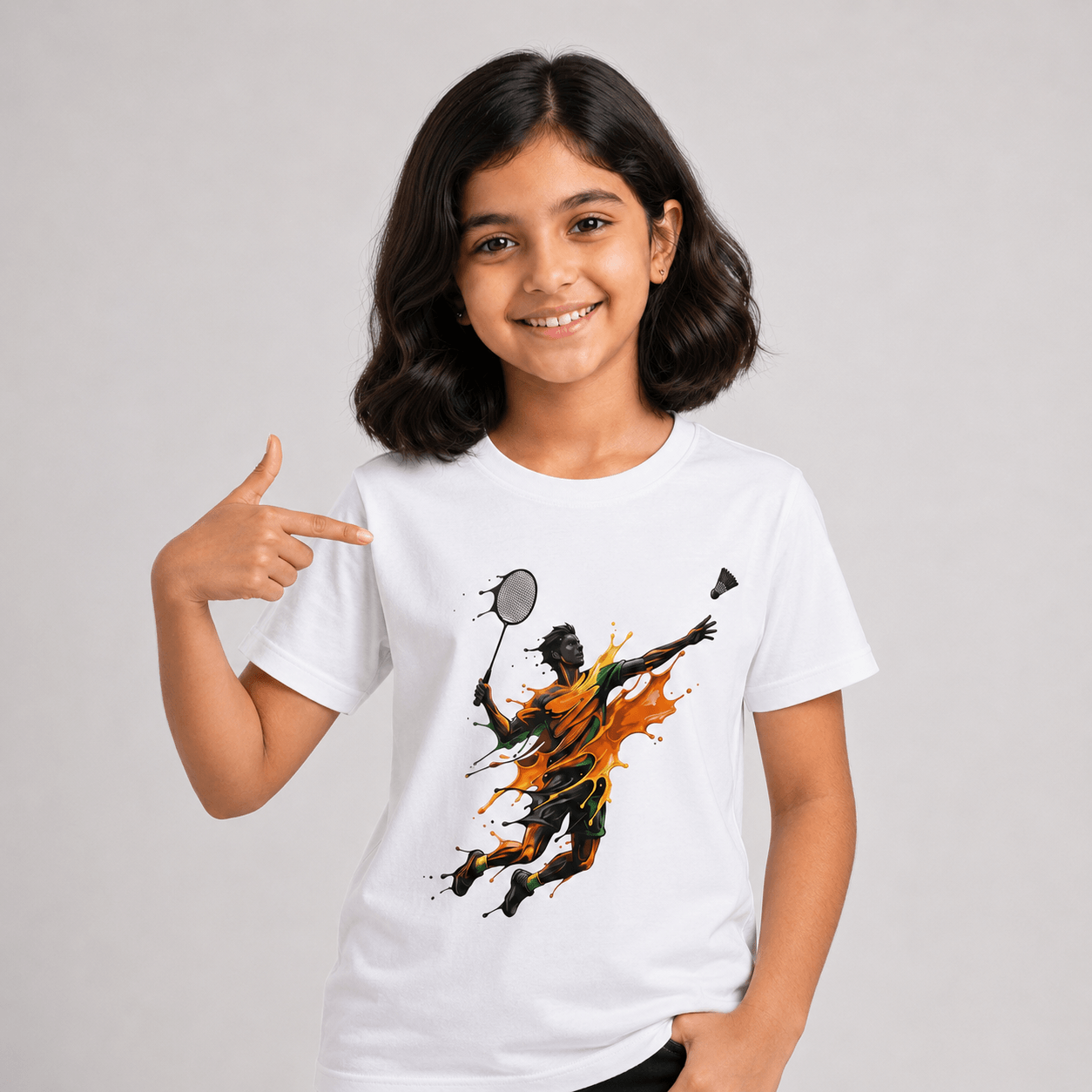 A young girl with dark hair smiling and pointing at a white graphic t-shirt. The t-shirt features a stylized print of a badminton player in black and orange paint splatter.