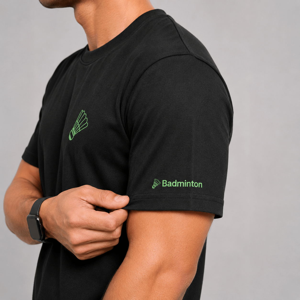 A man wearing a black short-sleeve t-shirt with a green shuttlecock logo on the left chest and 'Badminton' text with a shuttlecock logo on the right sleeve, shown against a neutral gray background.