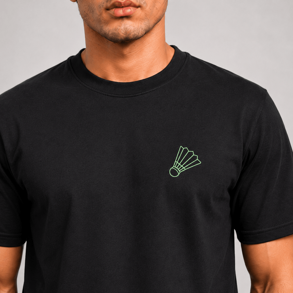 A man wearing a black short-sleeve crewneck t-shirt with a small green shuttlecock outline printed on the left chest, shown against a neutral gray background.