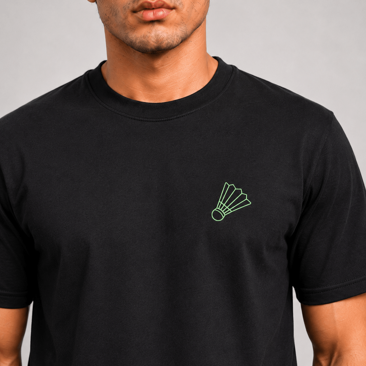 A man wearing a black short-sleeve crewneck t-shirt with a small green shuttlecock outline printed on the left chest, shown against a neutral gray background.