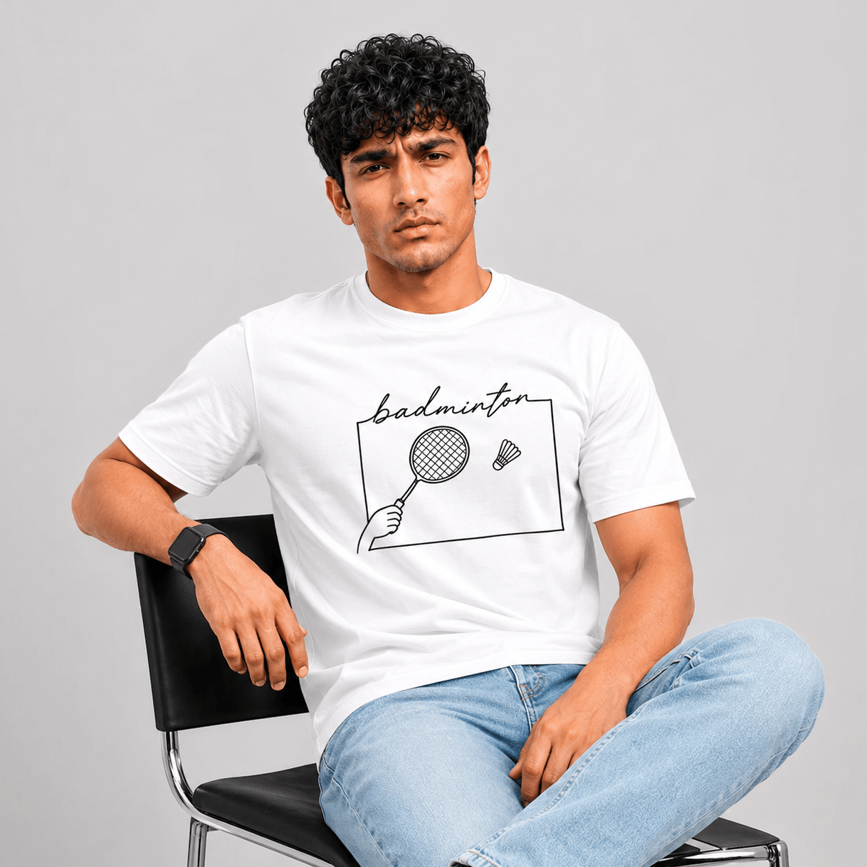 A man sitting on a black chair wearing a white graphic t-shirt with a black hand-drawn style 'badminton' design, paired with light blue jeans, against a solid gray studio background.
