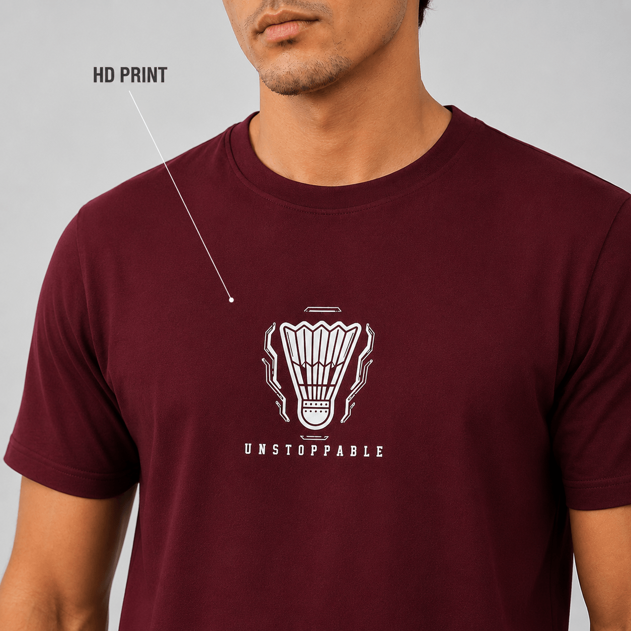 A man wearing a maroon short-sleeve t-shirt with a white graphic of a badminton shuttlecock and the word 'UNSTOPPABLE' printed on the chest. A line points to the graphic with the text 'HD PRINT' indicating a high-definition print feature.