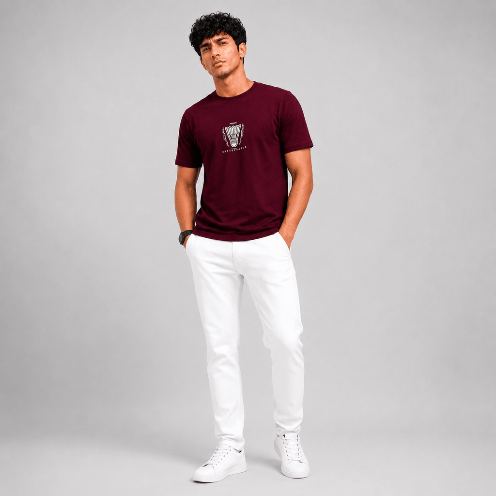 A man modeling a short-sleeve crewneck t-shirt in maroon with a small graphic on the chest, paired with white pants and white sneakers, standing against a light gray studio background.