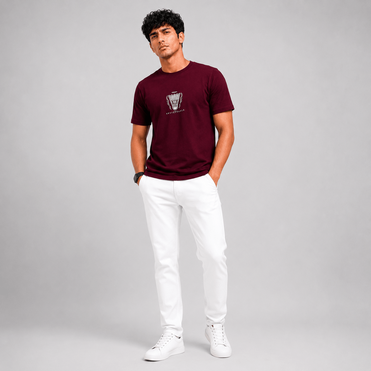 A man modeling a short-sleeve crewneck t-shirt in maroon with a small graphic on the chest, paired with white pants and white sneakers, standing against a light gray studio background.