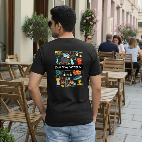 Man wearing a black t-shirt with colorful graphics and text, standing on a street with tables and chairs.