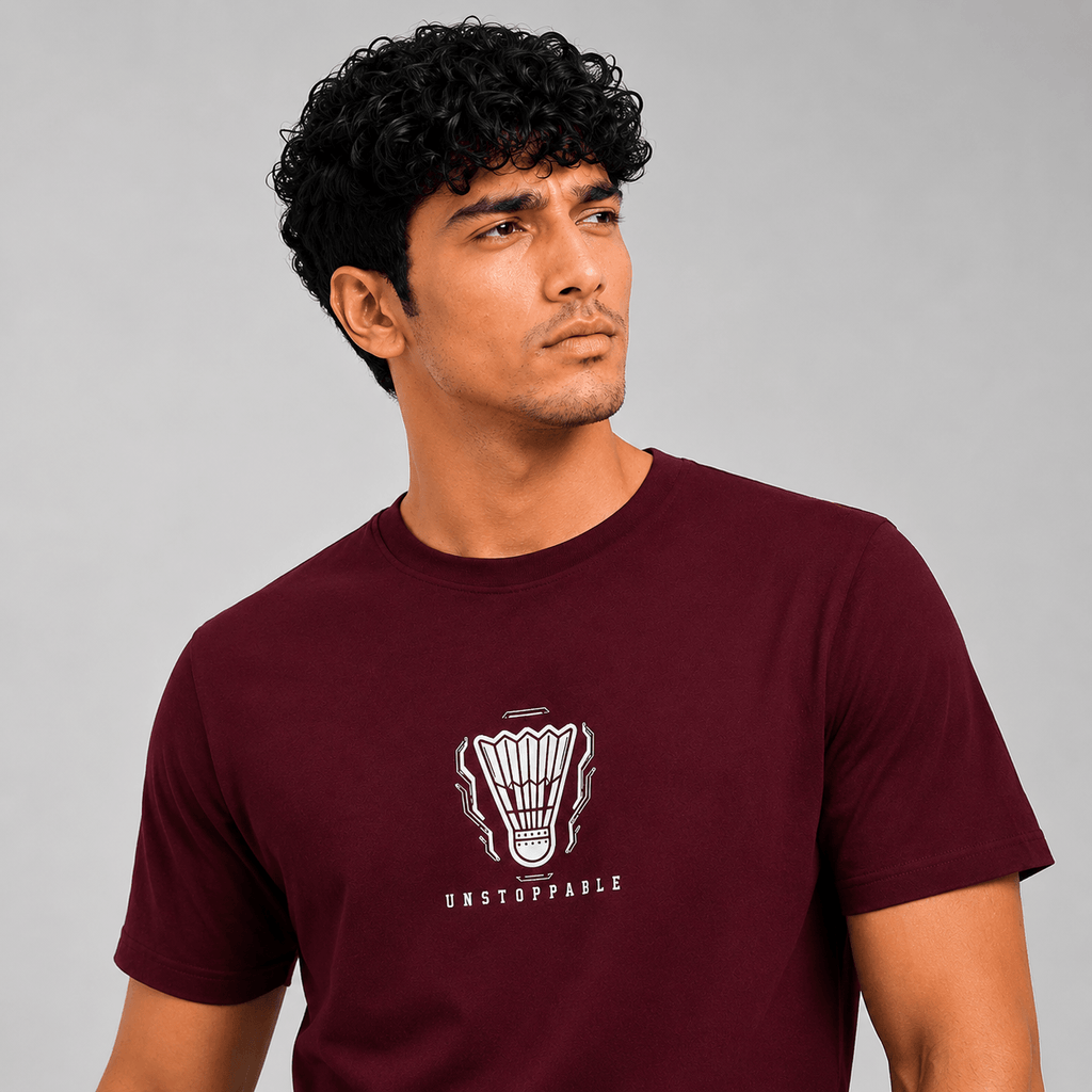 A man wearing a maroon short-sleeve t-shirt with a white shuttlecock graphic and the word 'UNSTOPPABLE' printed on the chest, set against a plain gray studio background.