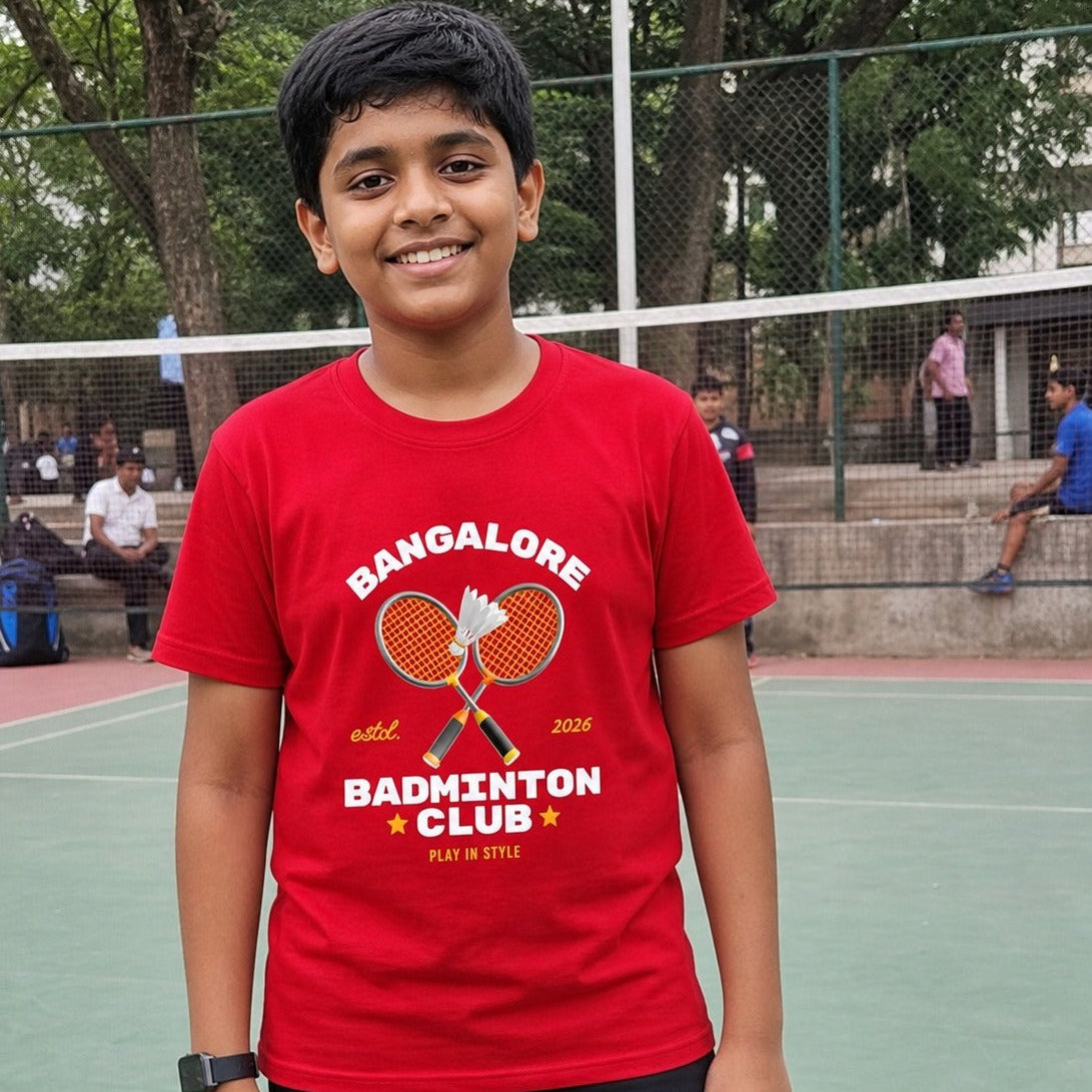 Young boy wearing a red t-shirt with 'Bangalore Badminton Club' print on a tennis court.
