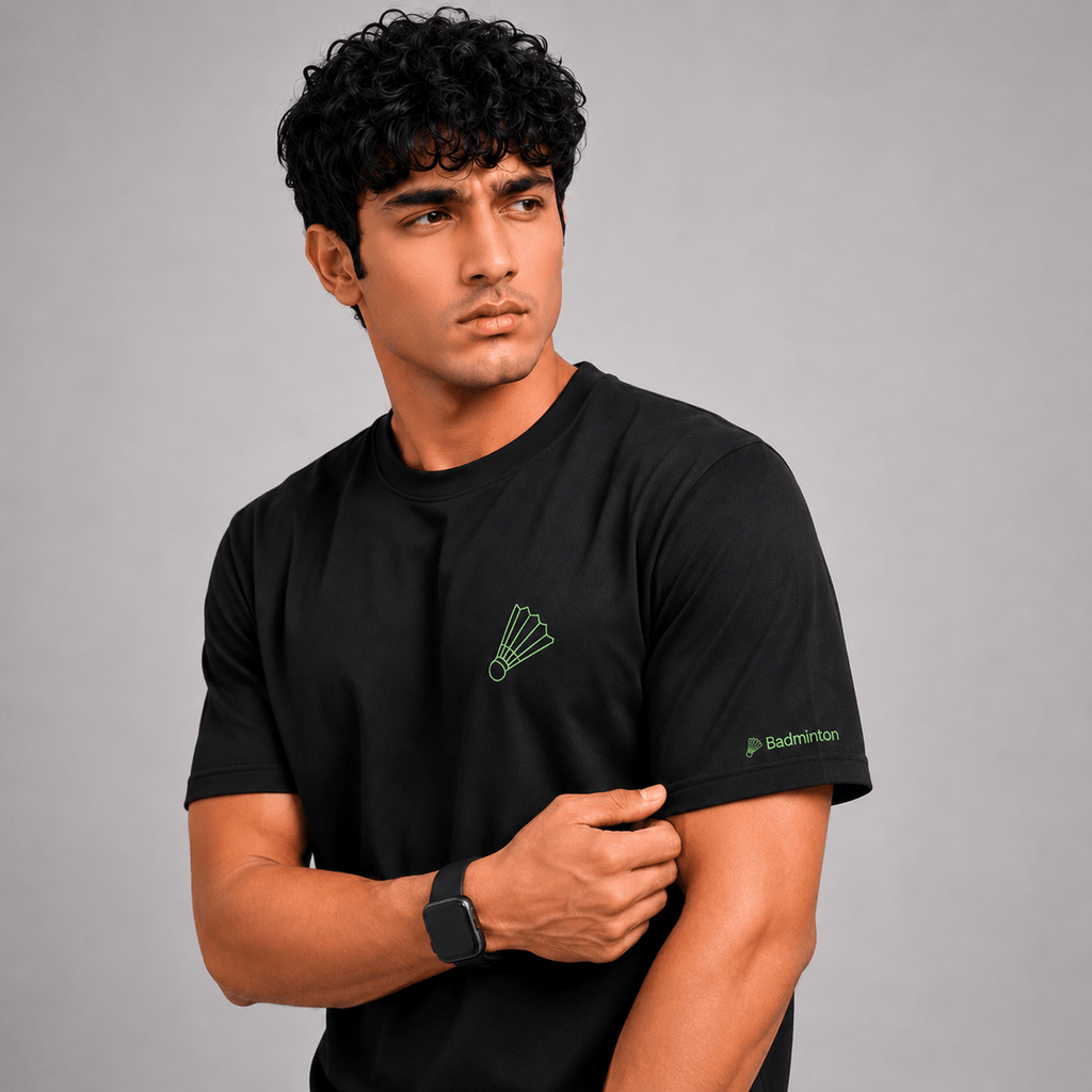 A man wearing a black short-sleeve athletic t-shirt with a green badminton racket logo on the left chest and 'Badminton' text on the right sleeve, posed against a solid gray background.
