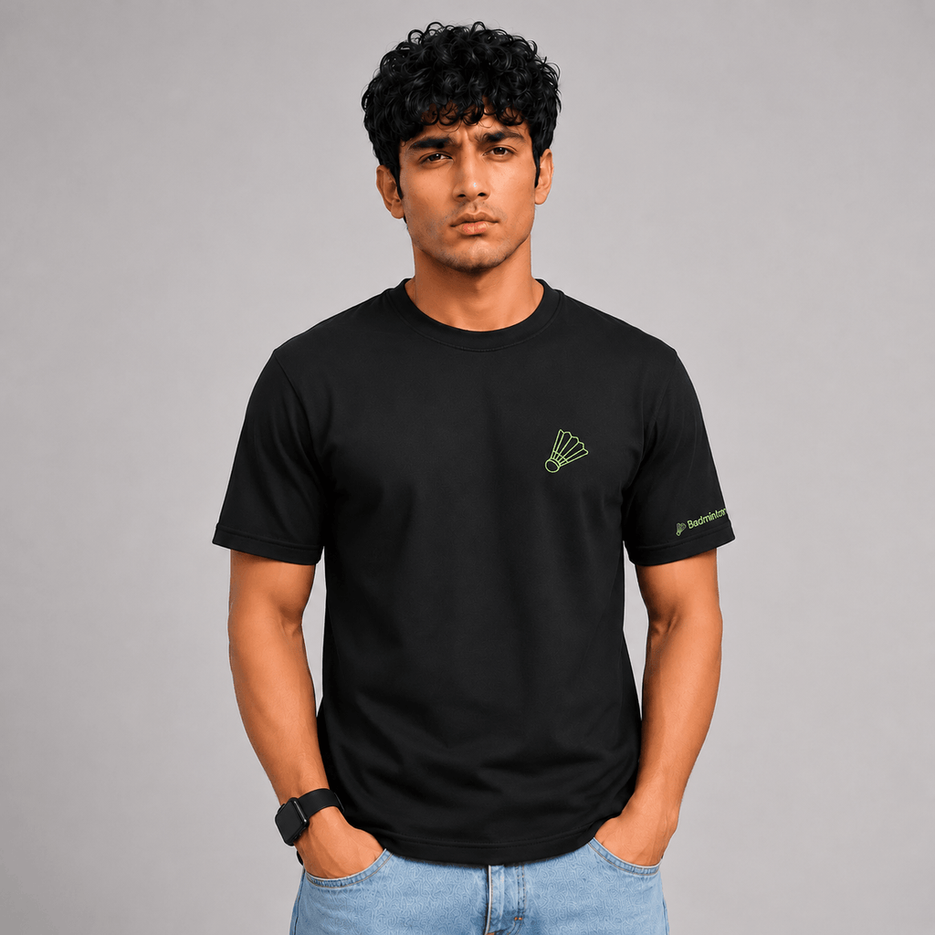 A man wearing a black short-sleeve crewneck t-shirt with a small green shuttlecock logo on the left chest and a green brand logo on the right sleeve, paired with light blue jeans, against a solid gray studio background.