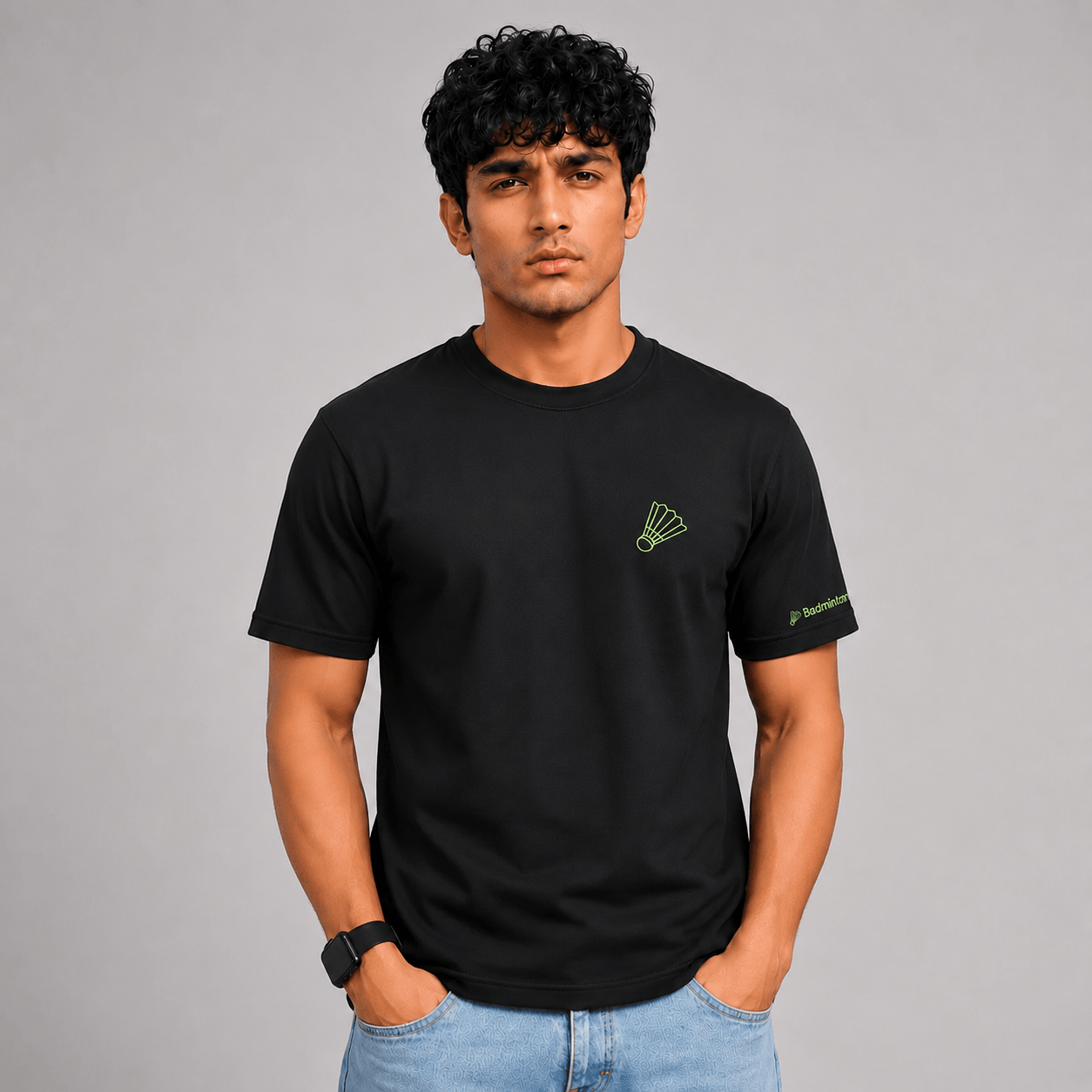 A man wearing a black short-sleeve crewneck t-shirt with a small green shuttlecock logo on the left chest and a green brand logo on the right sleeve, paired with light blue jeans, against a solid gray studio background.